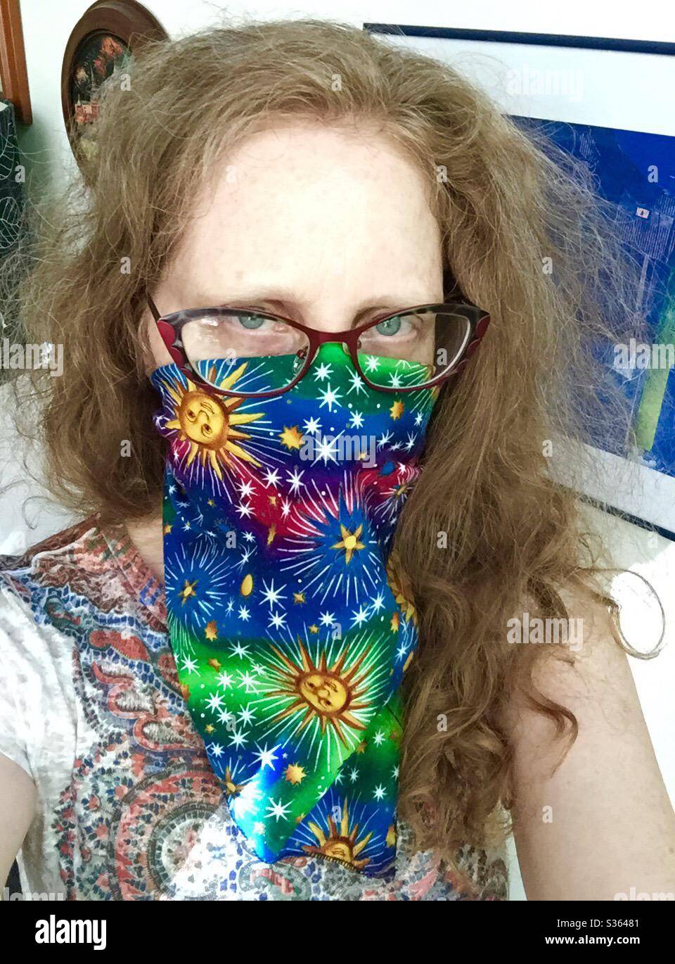 Mujer de pelo rojo con gafas usa un pañuelo de lujo con sol y estrellas como máscara, PPE, para protegerse contra la transmisión del virus COVID19. Foto de stock