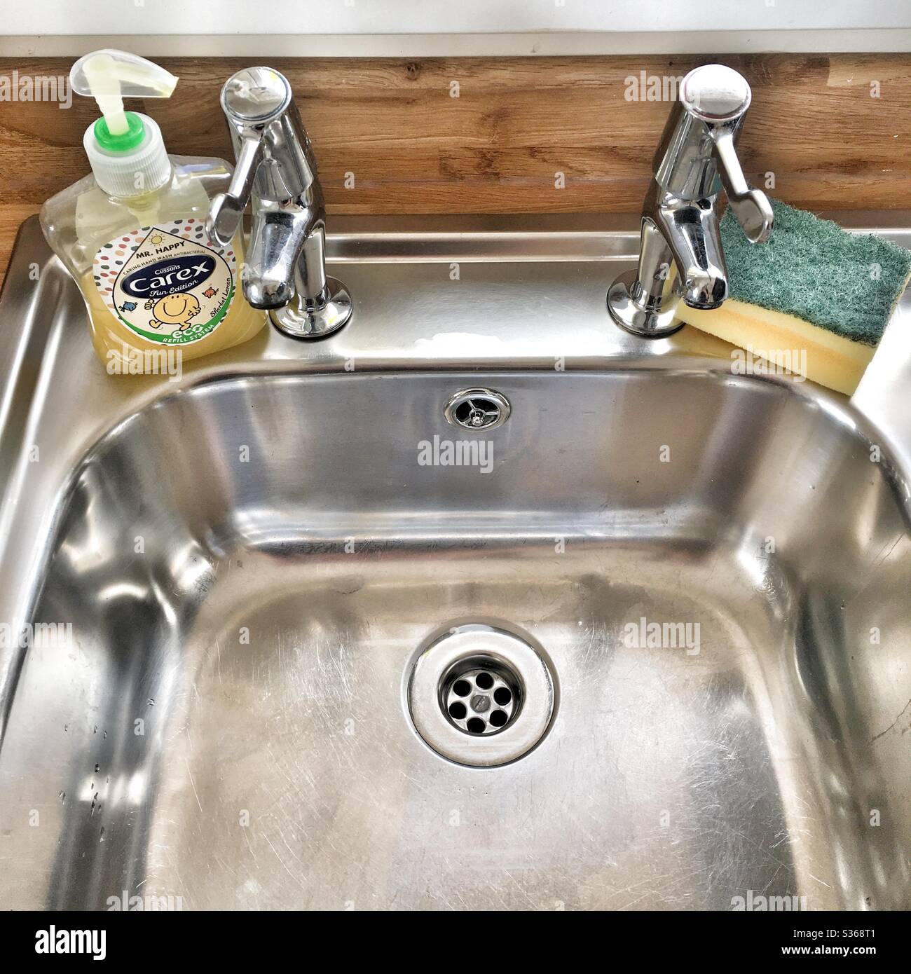 Una fotografía de un fregadero de acero inoxidable impecablemente limpio y grifos con una botella de jabón de manos Carex y una esponja. Limpieza, limpieza de casa, ocd, concepto de lavado de manos. Foto de stock
