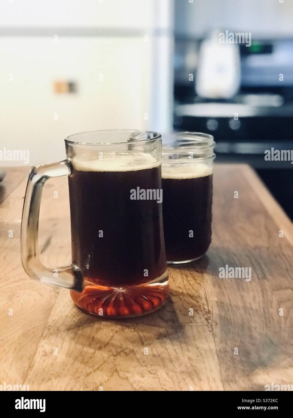Dos vasos de cerveza casera oscura se sientan en un mostrador de madera en una cocina. Foto de stock