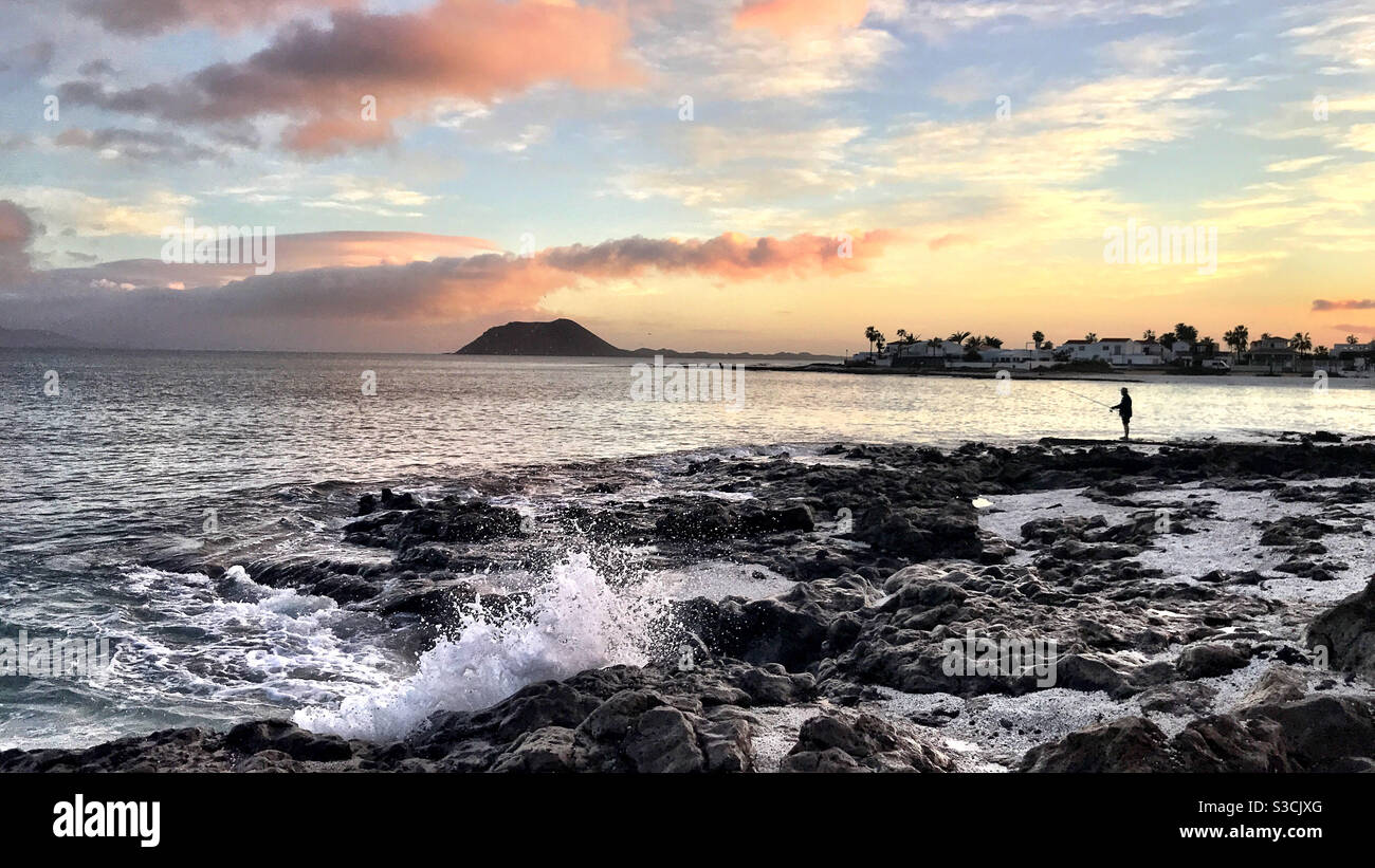 Pesca en línea al amanecer, playa de Corralejo, Fuerteventura, Islas Canarias. Hermosa vista, sol naciente, nubes rosadas, aguas tranquilas, olas rompiendo en rocas, vista hacia la pequeña isla de los Lobos Foto de stock