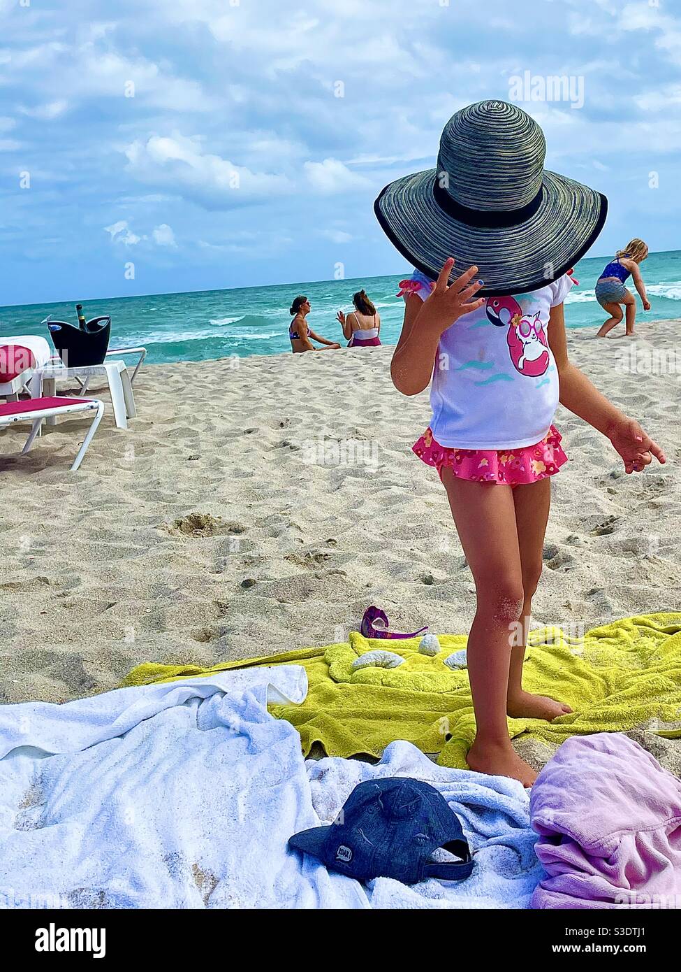 Una niña pequeña con traje de baño rosa y blanco se esconde bajo un sombrero  de sol adulto en un complejo de playa en Miami South Beach, Florida,  Estados Unidos Fotografía de