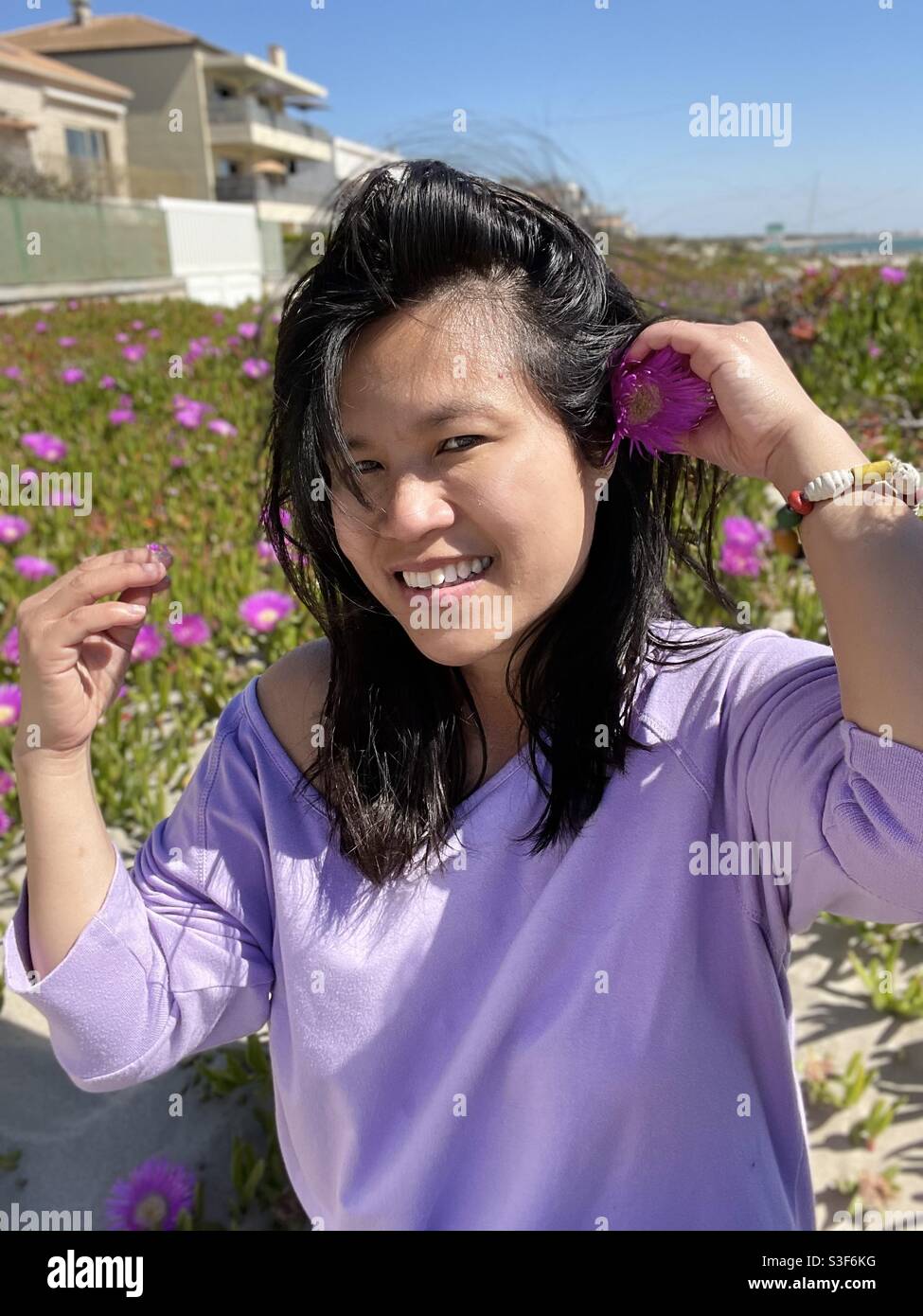 Exótica mujer filipina del sudeste asiático poniendo una flor púrpura en su largo cabello negro, frente a suculentas flores de plantas en la playa de Carnon plage, cerca de Montpellier, Occitanie, al sur de Francia Foto de stock