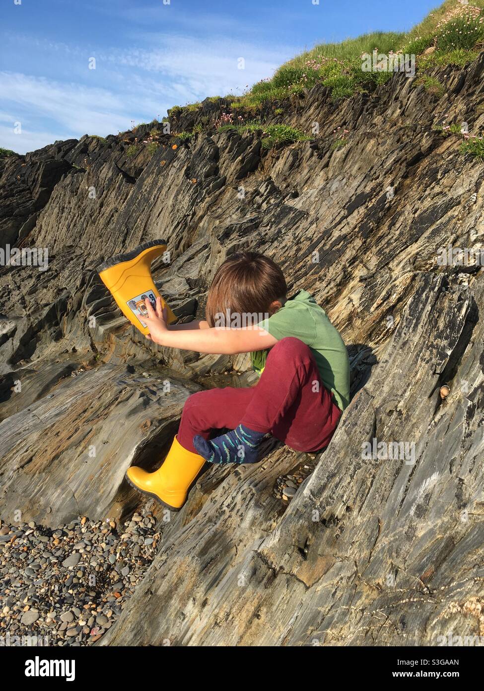 Niño vaciando piedras de la bota amarilla Wellington en la playa Foto de stock