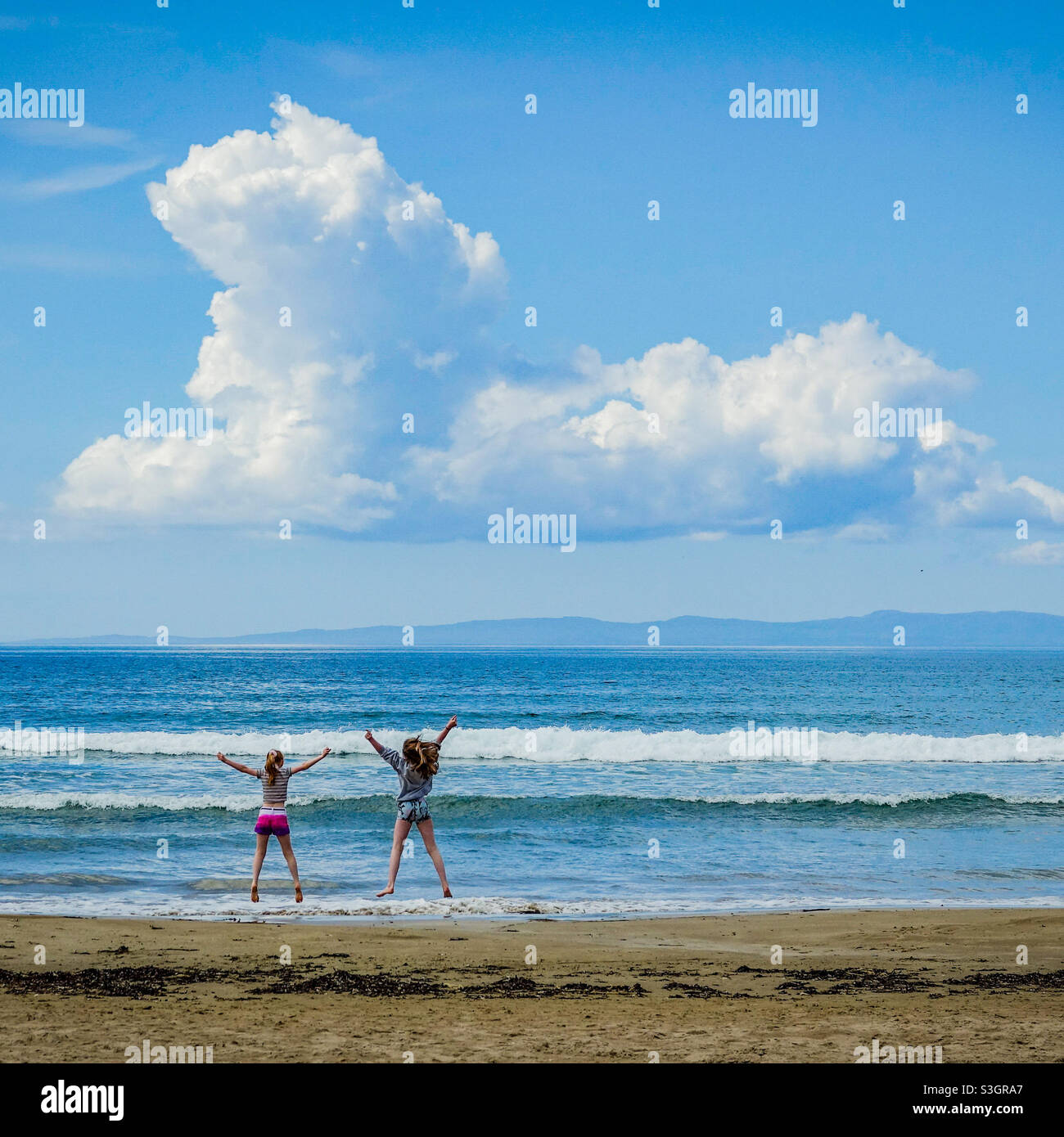 Dos chicas saltando en una playa, alcanzando una nube en forma de perro Foto de stock