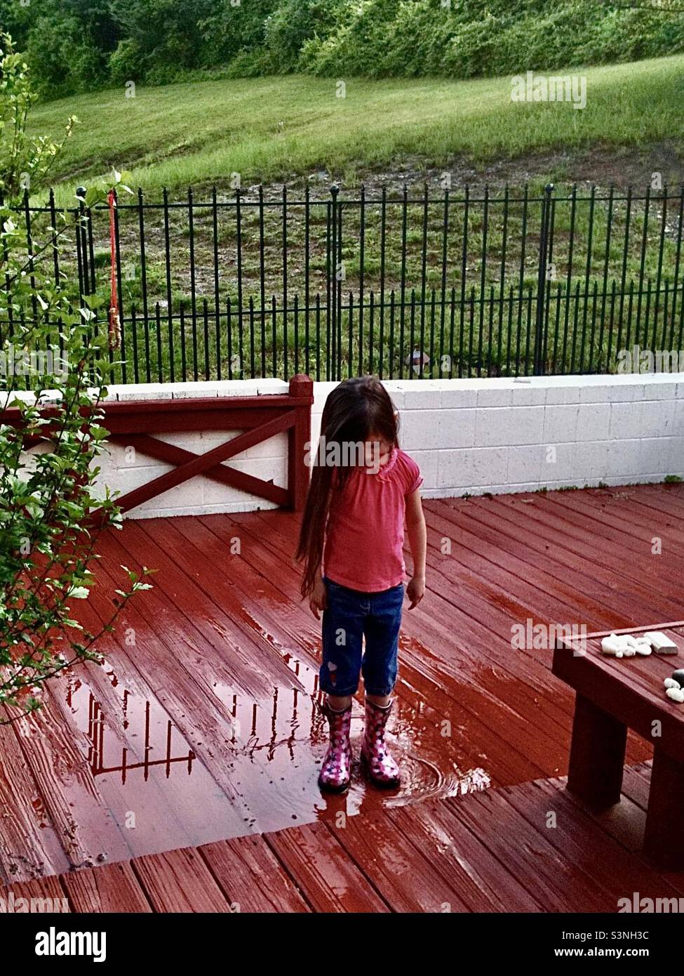 Una niña con el pelo largo y oscuro en botas de lluvia rosa después de una  tormenta de primavera mira hacia abajo a sus botas mientras chapoca en un  charco en una