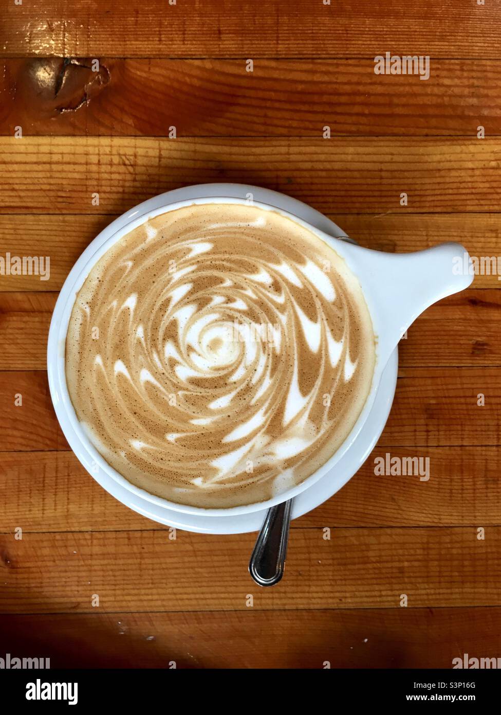 Café latte con hermoso patrón de espuma en tapa blanca con cuchara sobre mesa de madera marrón Foto de stock