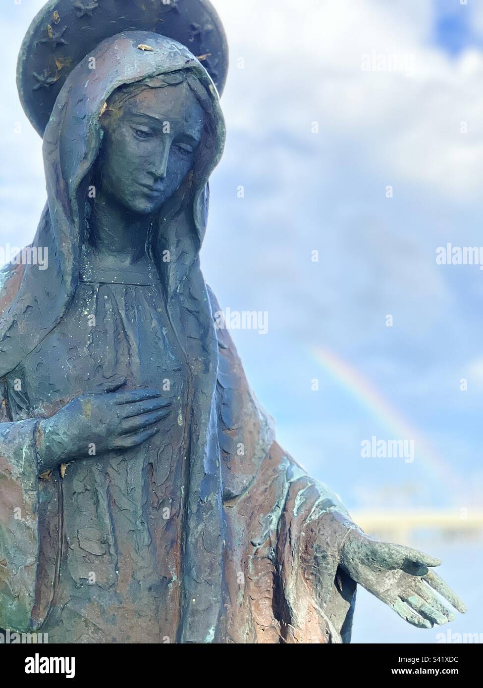 Retrato, telón de fondo del arco iris, estatua de bronce de la Virgen María, coronada con 12 estrellas, mano derecha sobre el corazón, mano izquierda extendida para ayudar, visión de ángulo bajo, mirando en la cara de María Foto de stock