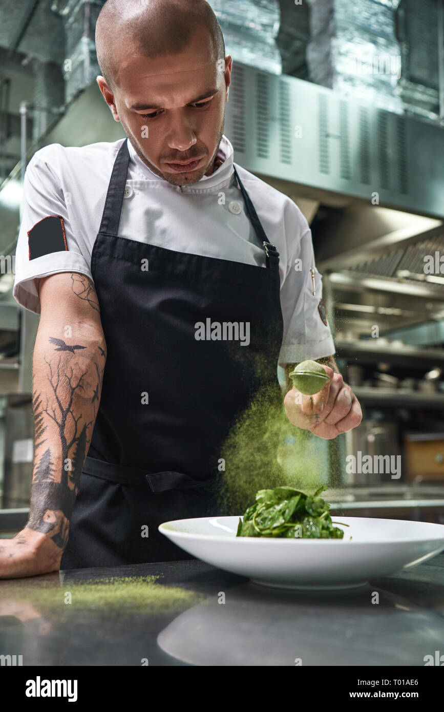 Comer sano. Vertical de hermoso macho chef en delantal negro agregando especias en ensalada mientras está de pie en la cocina de un restaurante. Foto de stock