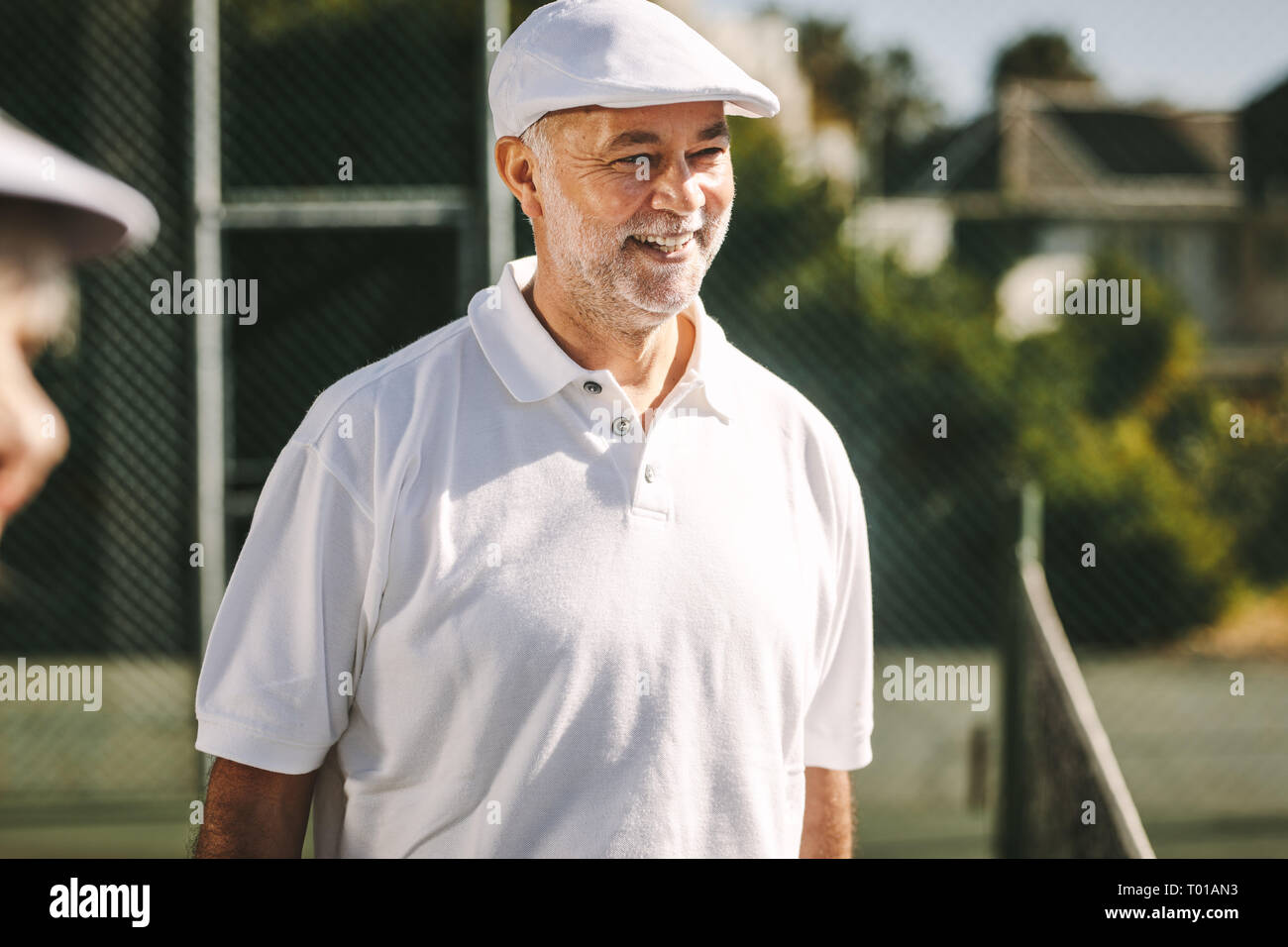 Senior alegre hombre de pie sobre una pista de tenis en un día soleado. Hombre sonriente en blanco camiseta de tenis y tapa de pie afuera. Foto de stock