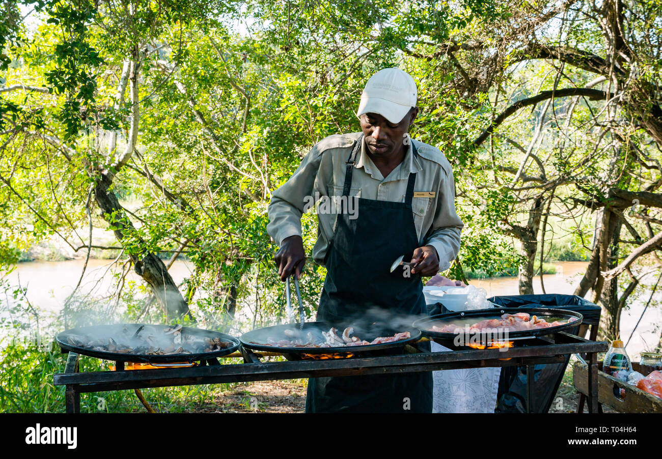 Hombre Negro cocinar el desayuno en la parrilla al aire libre con bacon y salchichas para el desayuno para los huéspedes de Safari, Sabi Sands Game Reserve, Sudáfrica Foto de stock