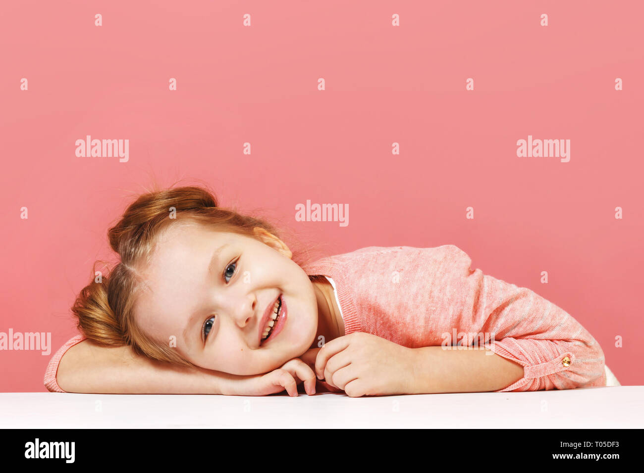 Closeup retrato de una linda niña con cabello bollos sobre fondo de color rosa. Sonriente niño preescolar reclinó su cabeza sobre la mesa Foto de stock