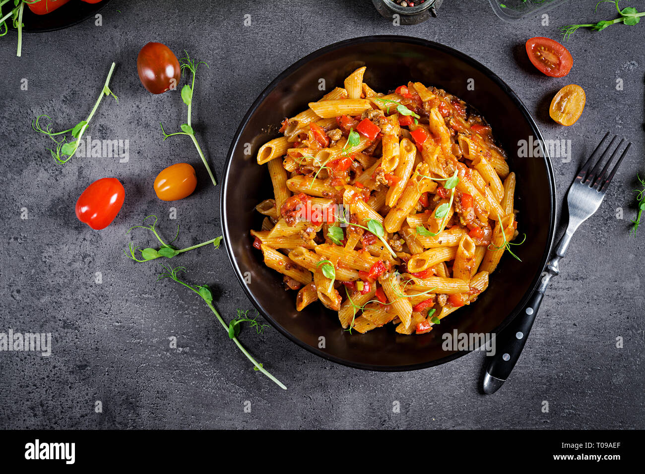 Pasta Penne en salsa de tomate con carne, tomates decorado con brotes de guisantes en una oscura mesa. Vista superior Foto de stock