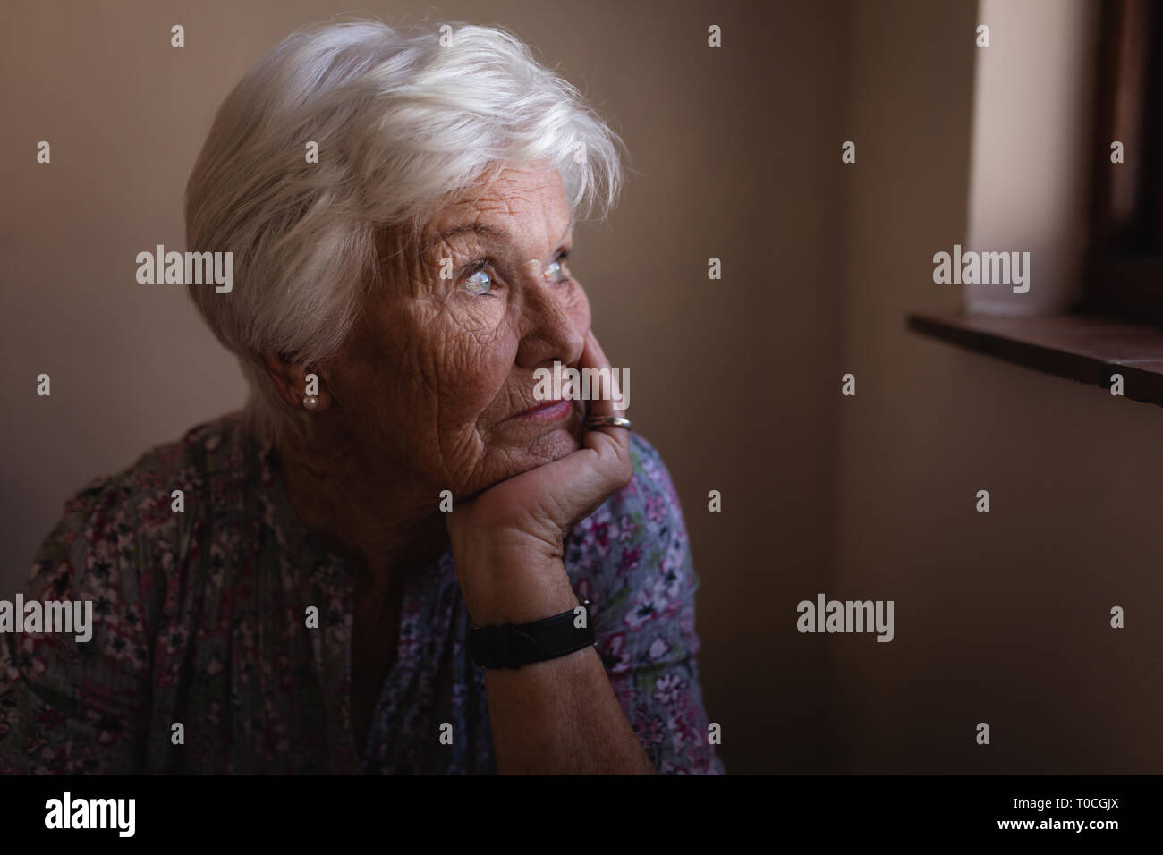 Pensativo activa mujer senior con la mano en el mentón mirando a través de la ventana de la cocina de casa Foto de stock