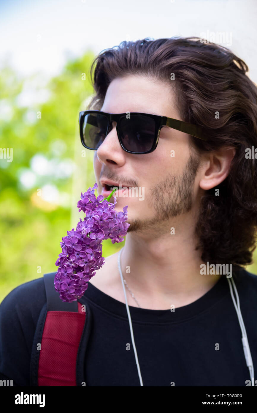 Hombre joven morenita con mucho pelo rizado y gafas de sol tiene flores frescas en su boca en una rama lila y mira hacia otro lado sobre un fondo difuminado. Foto de stock