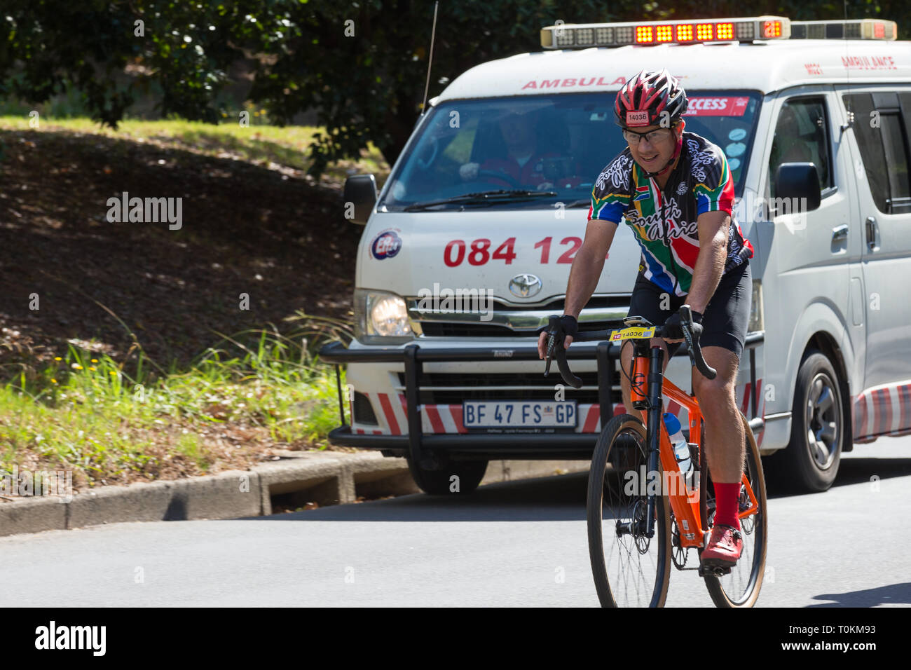 Ciclista vistiendo la camiseta de Sudáfrica y andar en bicicleta en una  carretera en una carrera ciclista, mientras que una ambulancia le va a  transferir el concepto de asistencia en carretera en
