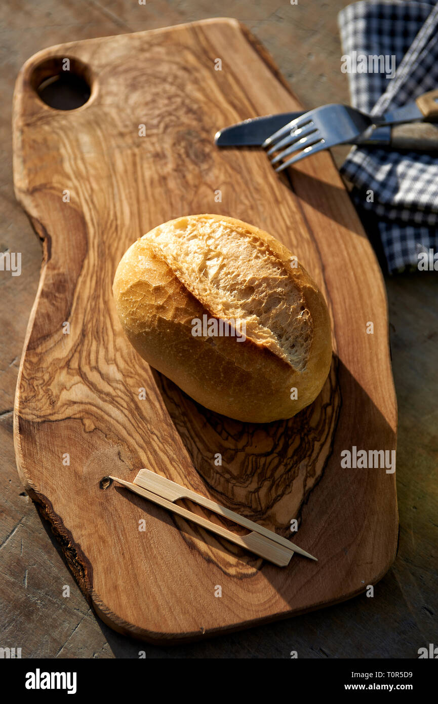 Ein Weizenbroetchen liegt auf einem Holzbrettchen und wartet darauf belegt zu werden. Als Deko liegt Messer und Gabel daneben, sowie Holzspiesschen Foto de stock