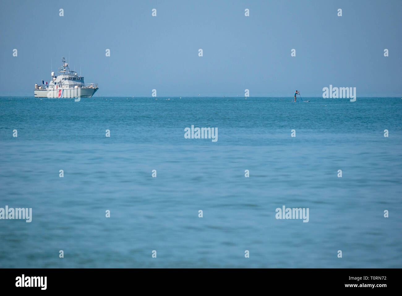 Varón soltero en un stand-up paddle board ligeramente a la costa de la playa en el norte de Francia con un barco de aduanas supervisan Foto de stock