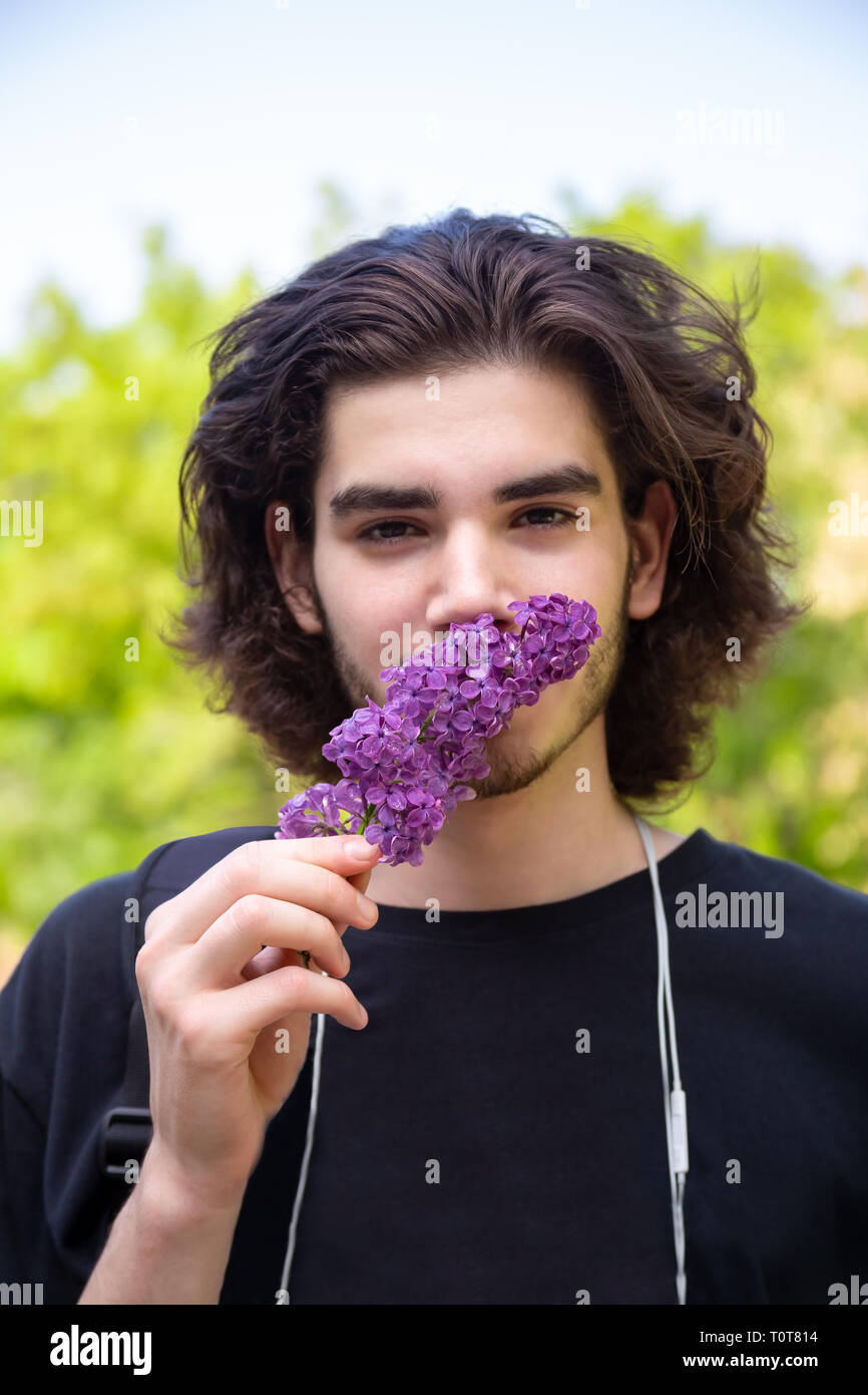 Joven morenita guy oliendo flores frescas en rama lila y mirando a la cámara de fondo borroso Foto de stock