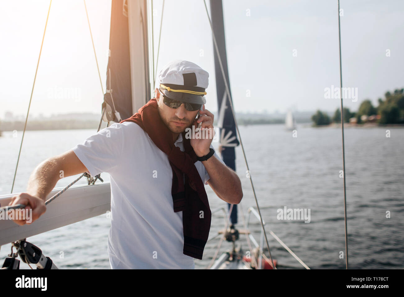 Serio y concentrado es capitán de yate y apoyarse sobre la baranda. Él sujete el teléfono en la mano y mira hacia abajo. Guy funciona. Itis sunset fuera. Foto de stock