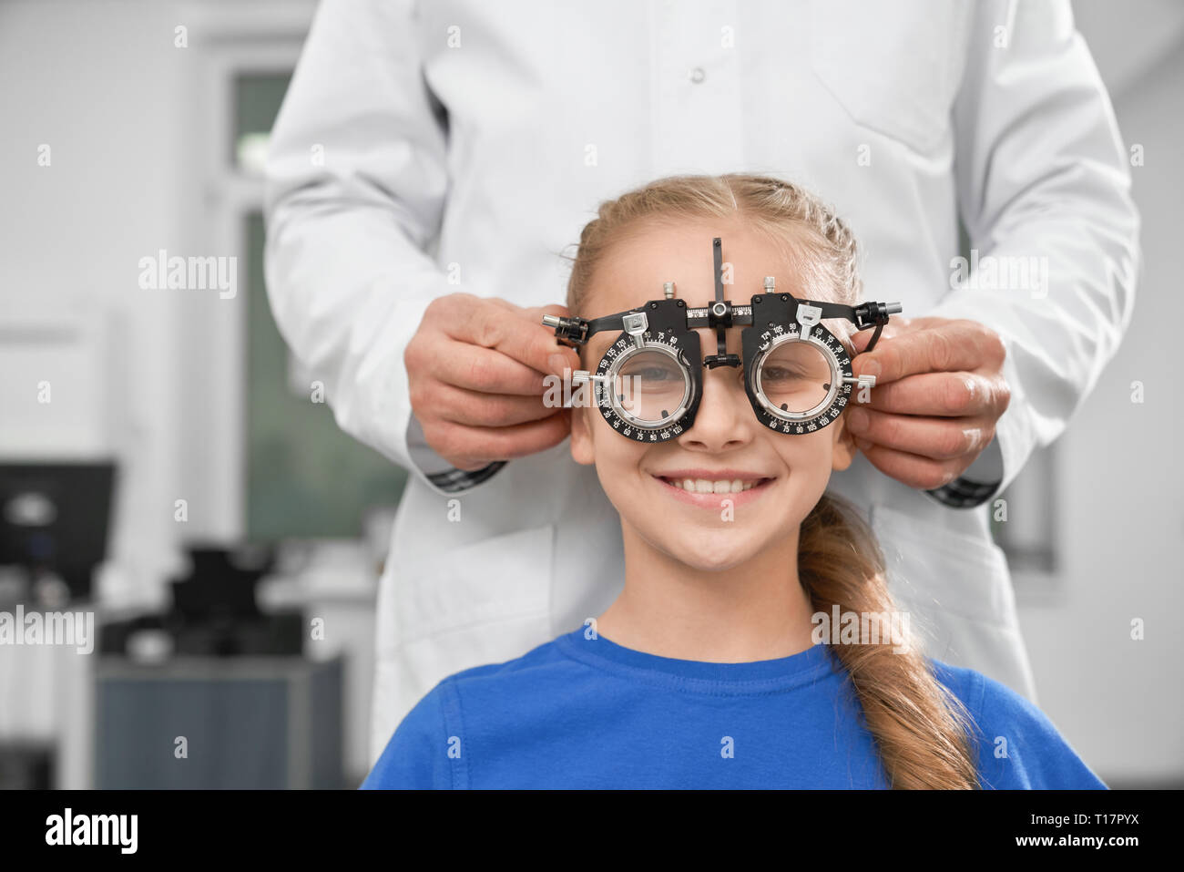 Oftalmólogo en bata blanca con equipo médico especial para la salud de los ojos, la comprobación de la visión. Sonriente chica sentada y mirando a la cámara a través de pruebas de gafas. Foto de stock