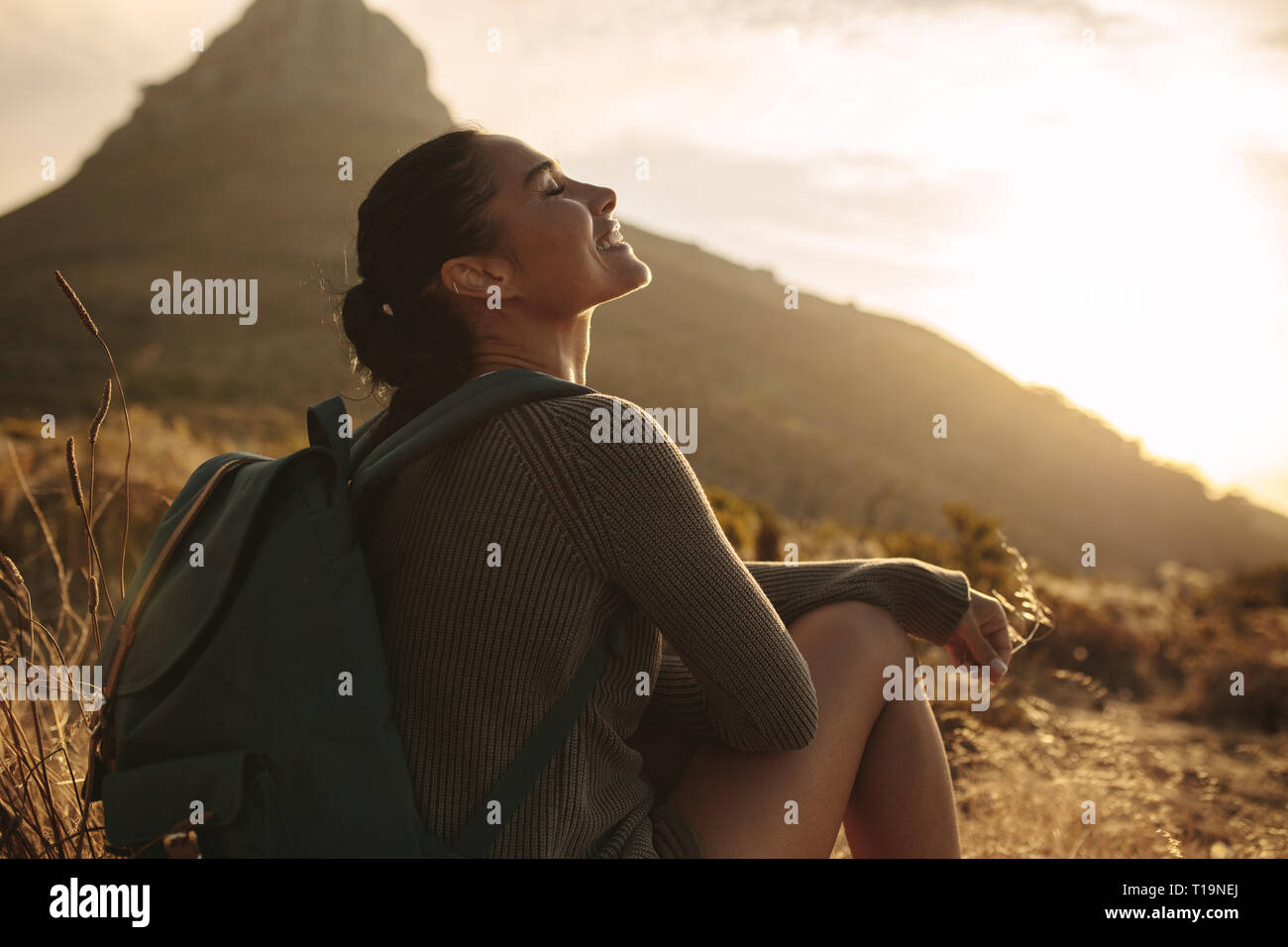 Mujer sentada en el suelo sonriendo con los ojos cerrados. Caucasian mujeres turistas tomando un descanso después de hacer senderismo en el país trail. Foto de stock