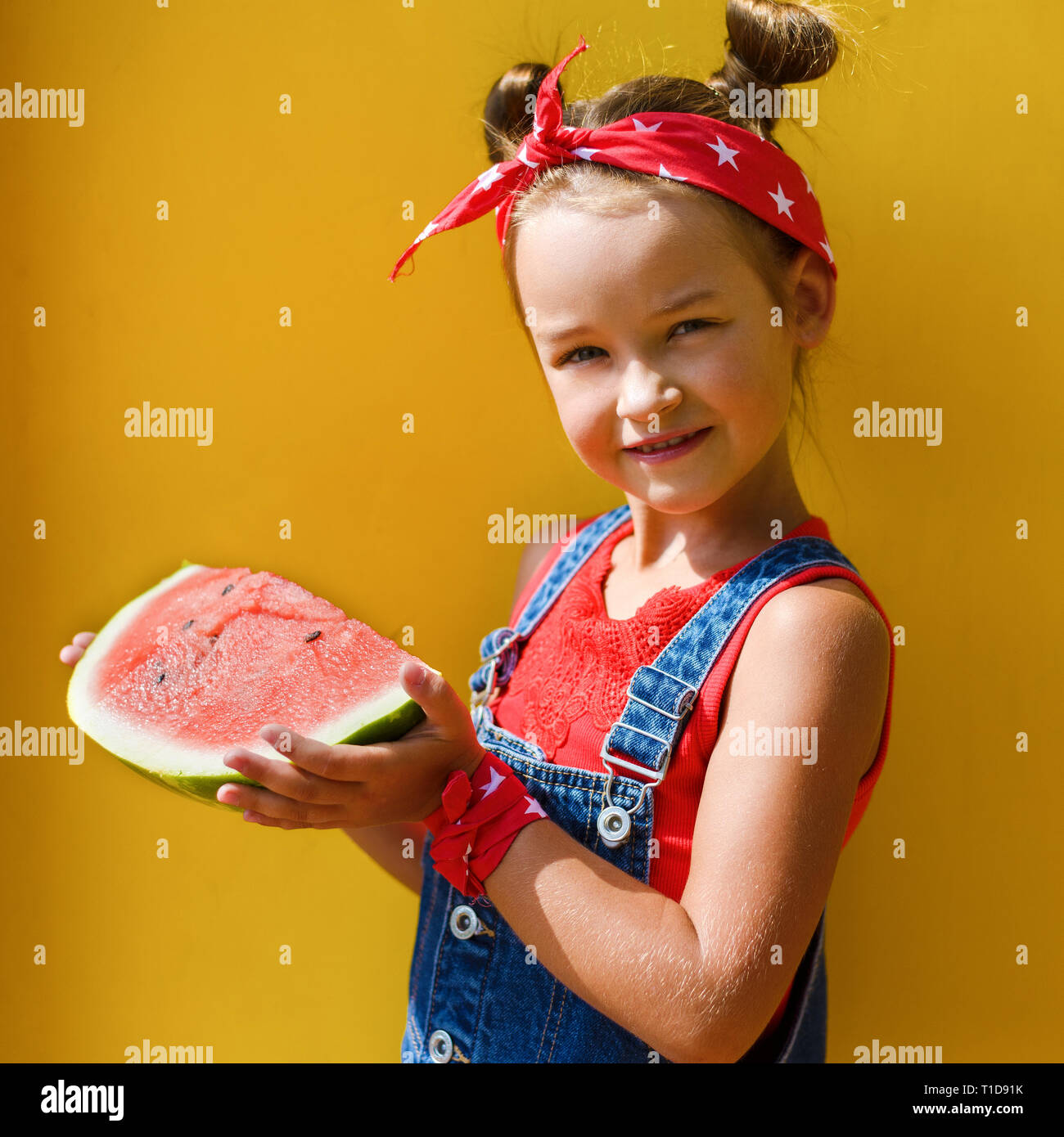 Retrato de sonrisa niña con colas de cabello en la cabeza en la camiseta  roja y pañuelo en la cabeza en un mono denim la celebración de sandía  madura. fondo amarillo Fotografía