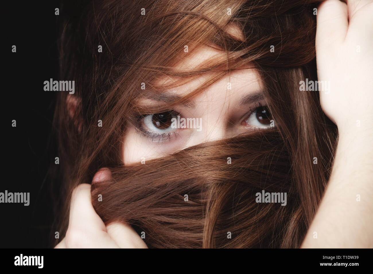 Soledad, estrés y ansiedad mujeres concepto. Closeup retrato Mujer hermosa morenaza, jovencita que cubren cara por largos pelos marrones sobre negro Foto de stock