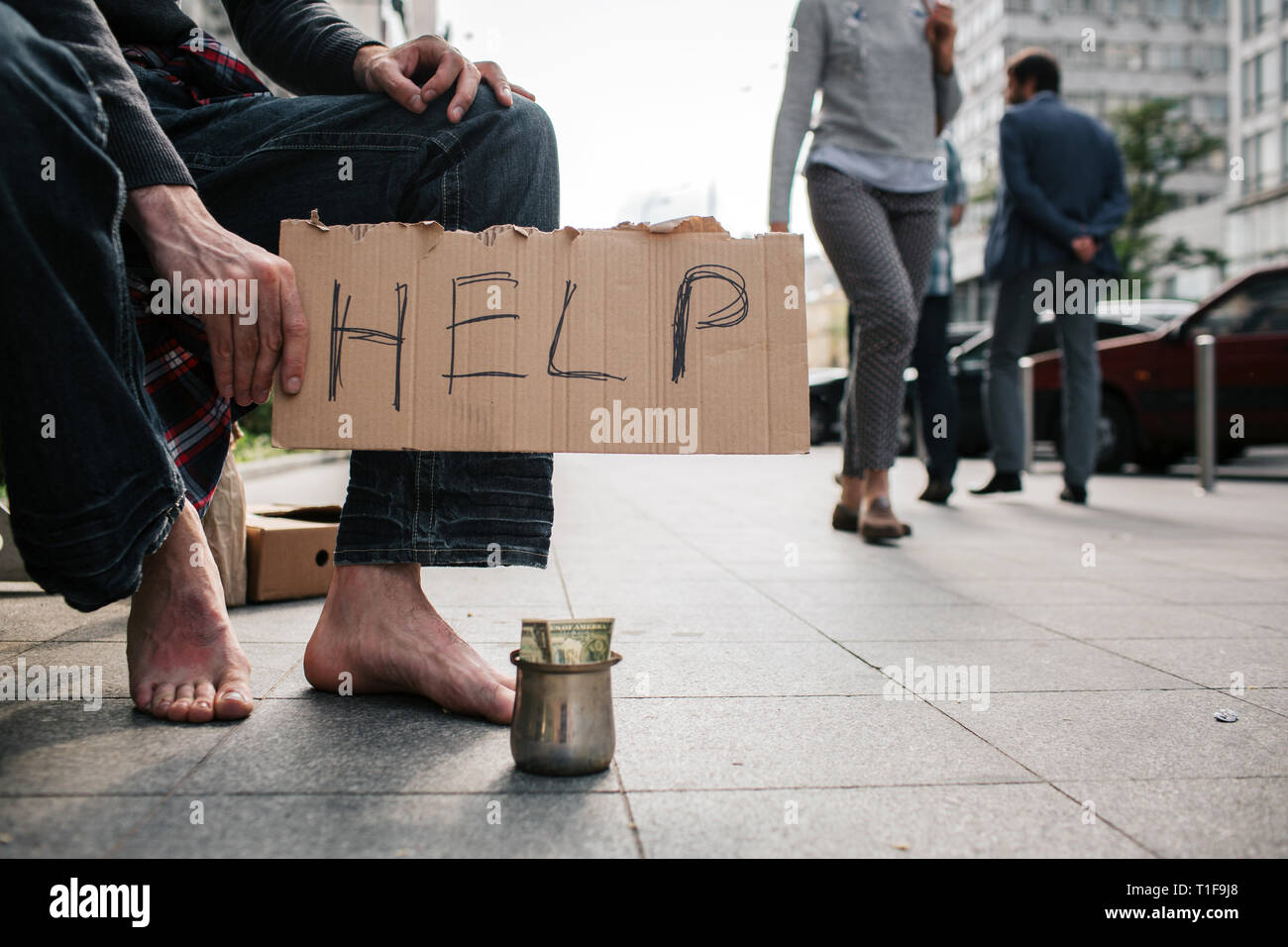 Una imagen del hombre sin piernas zapatos de pie en el suelo de hormigón. Existe una copa metálica con dinero en ella. Guy está sosteniendo un cartón que dice Foto de stock