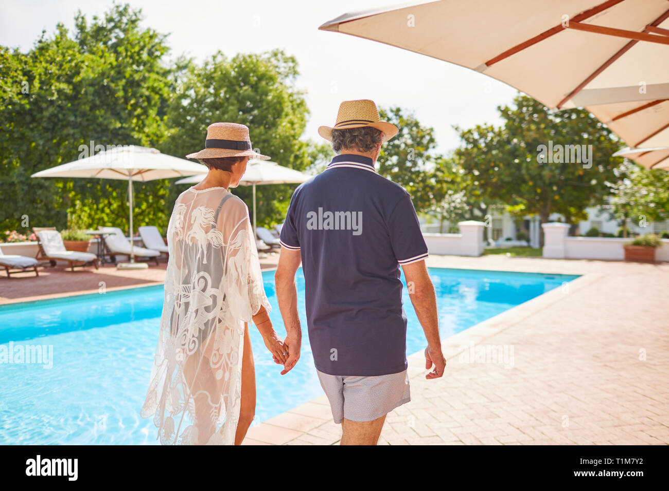 Pareja tomados de las manos, caminando junto a soleado complejo turístico piscina Foto de stock
