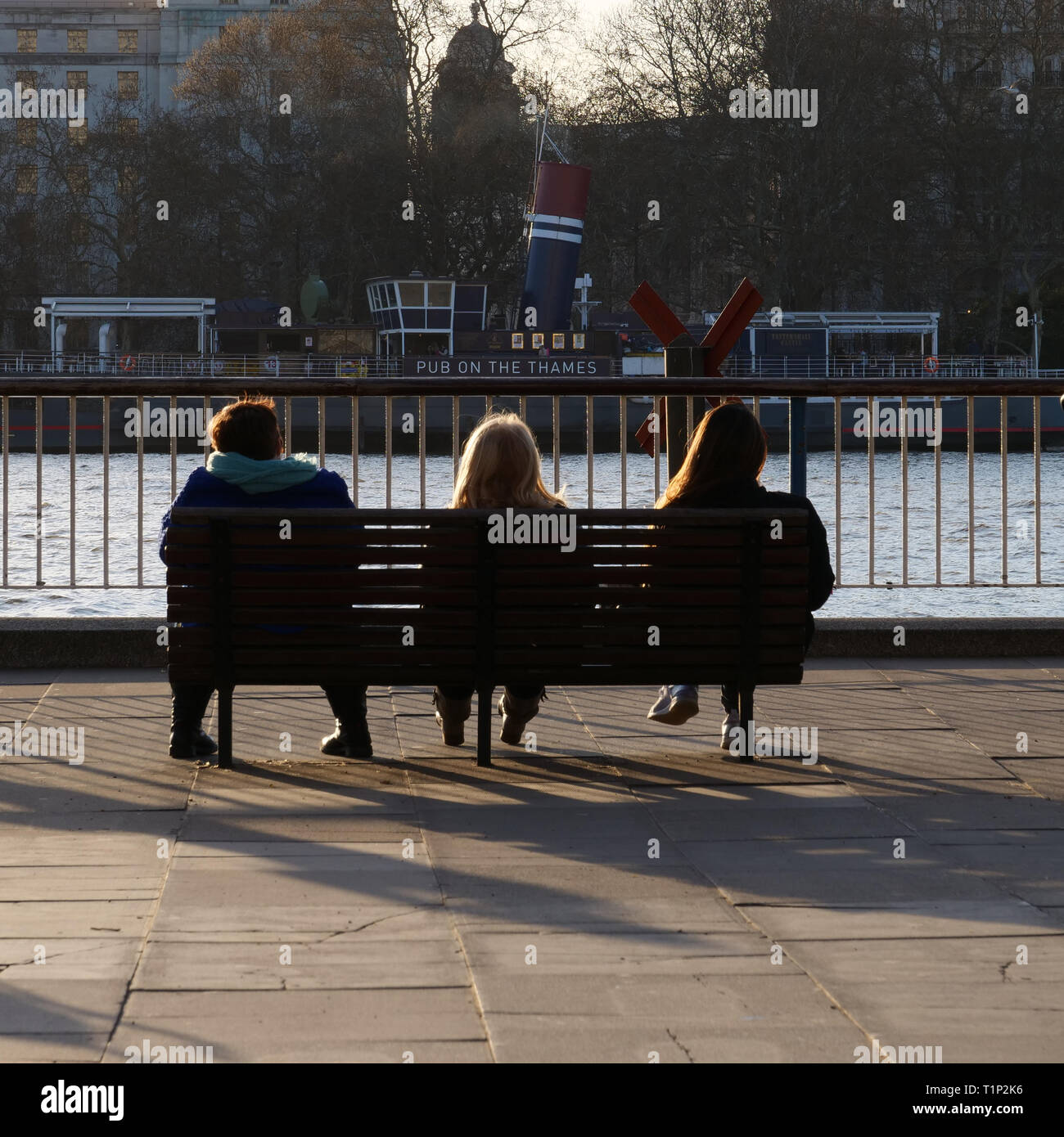 Tres personas sentadas en un banco en la orilla sur, con vistas sobre el Río Támesis en Londres, Reino Unido, y disfrutar del sol de la tarde en un cálido día de finales de marzo. Foto de stock