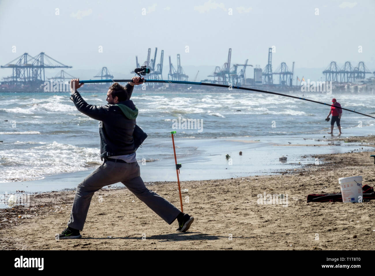 Valencia Vista al mar España Mar UN pescador en la playa lanza una caña de pescar al mar. Pesca, puerto de Valencia con grullas en el fondo Foto de stock