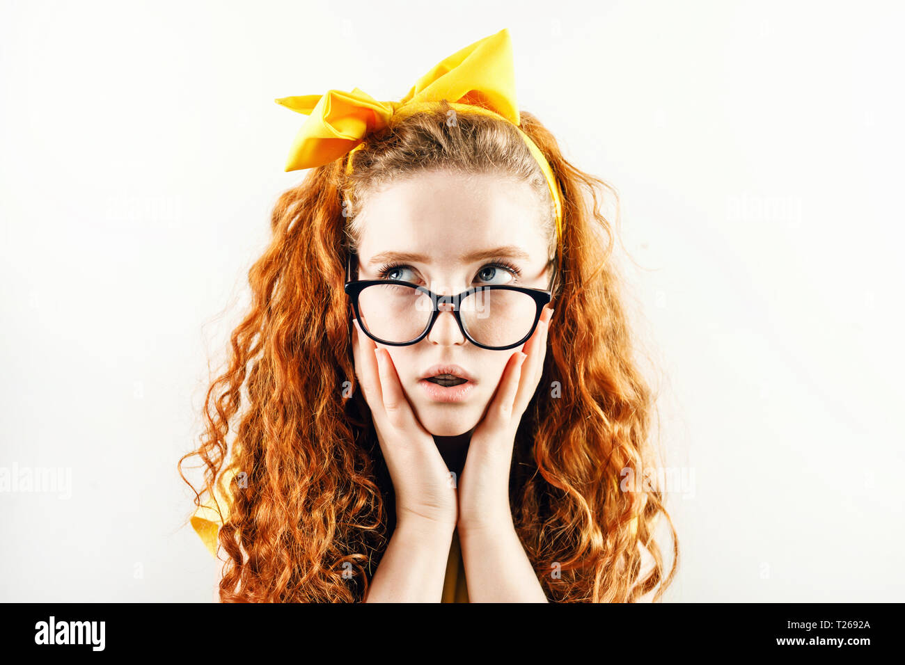 Schocked chica pelirroja rizada en los vasos con un arco amarillo sobre su cabeza vistiendo camiseta amarilla sosteniendo su hads cerca de su cara y apartar la mirada. Foto de stock