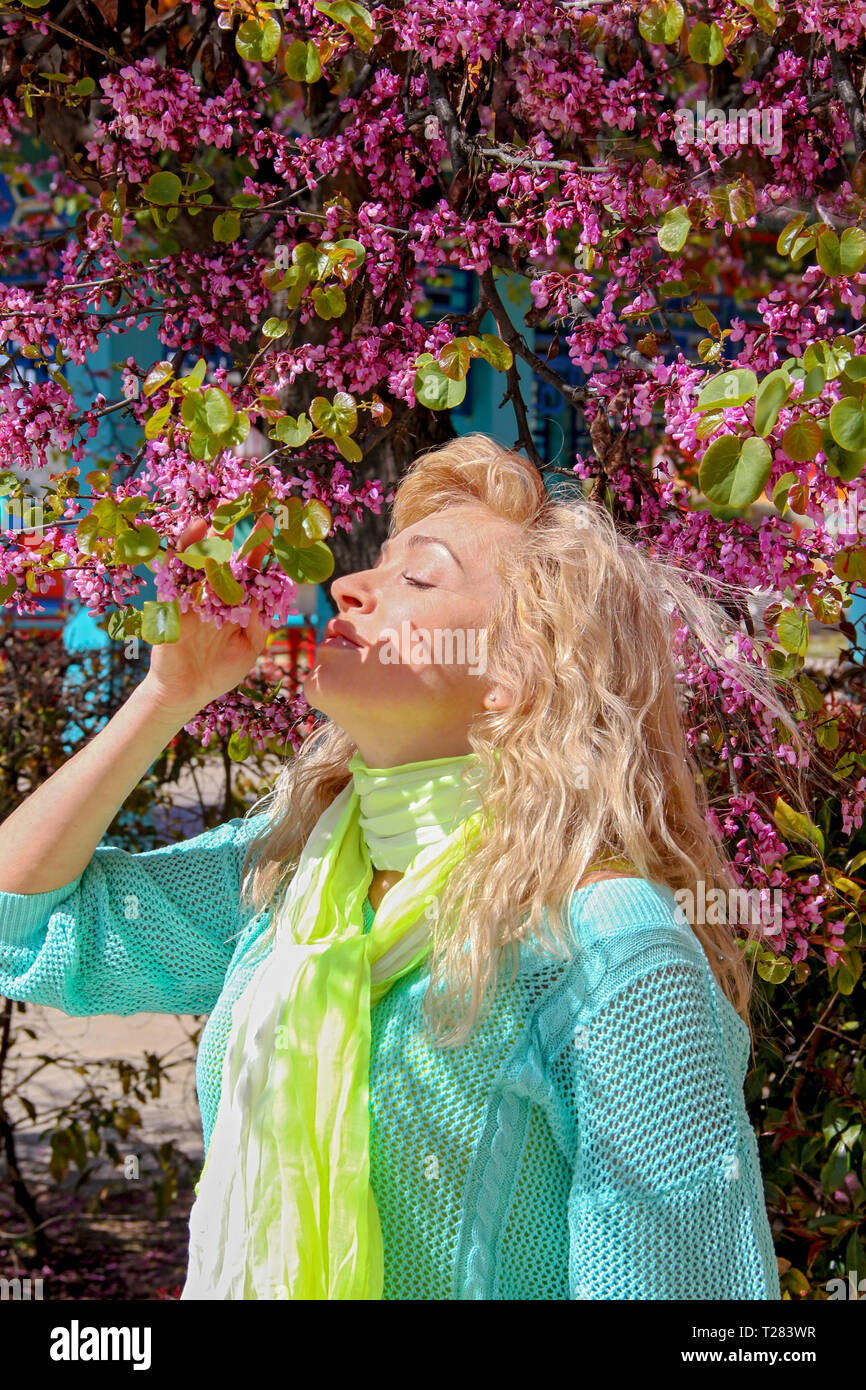 Una joven y bella mujer rubia con el pelo rizado está cerca de un árbol en flor en un cálido día de primavera o verano y huele flores púrpura con los ojos cerrados. Copie el espacio para su inscripción. Foto de stock