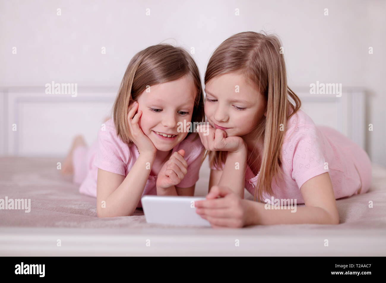 Dos niñas (hermanas) en pijama rosa en la cama viendo un smartphone y leer un e-book. El enfoque selectivo. Foto de stock