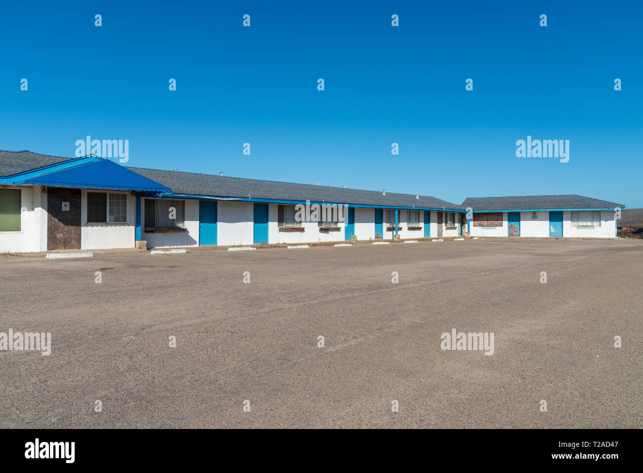 Amplia visión del abandono de motel con ribete azul y paredes blancas bajo el brillante cielo azul. Estacionamiento pavimentado. Foto de stock