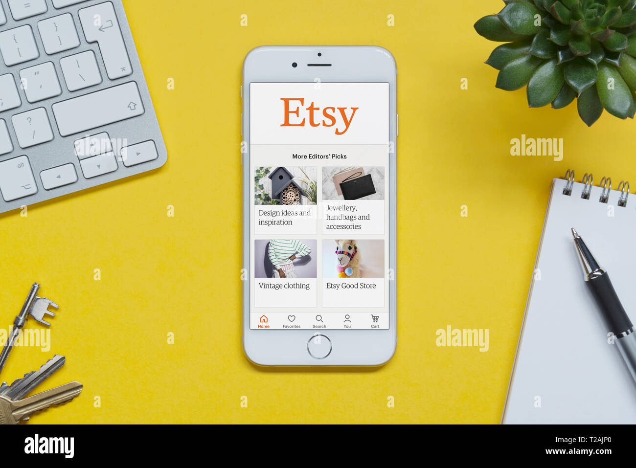 Un iPhone que muestra el sitio web Etsy descansa sobre un fondo amarillo tabla con un teclado, teclas, bloc de notas y Planta (uso Editorial solamente). Foto de stock