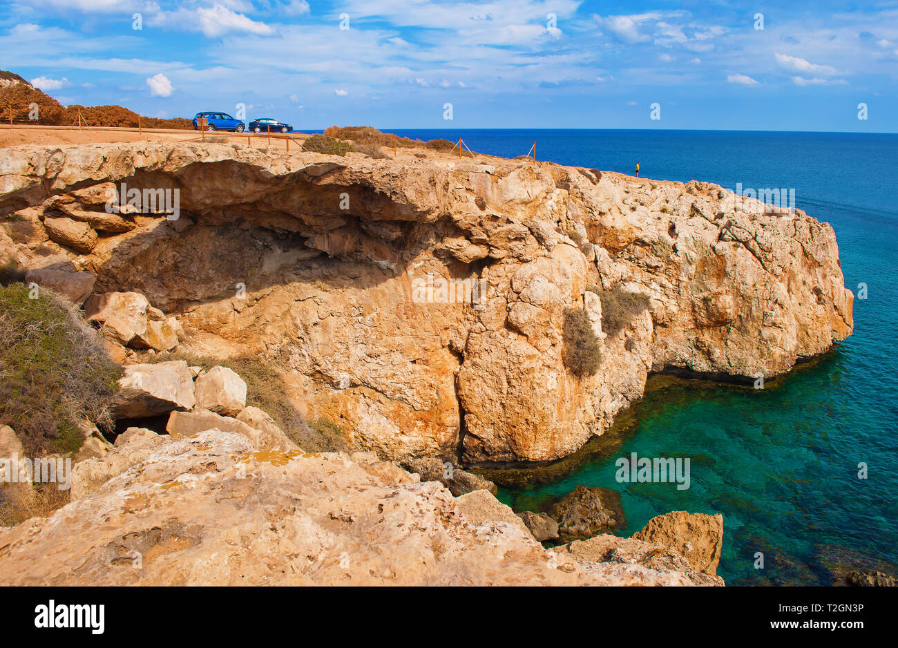 Una pequeña figura de hombre de pie en el borde de un acantilado. Dos autos cerca de una bahía cerca de Cape Greco, Chipre. Cerca de la costa de roca verde profundo azul transparente Foto de stock
