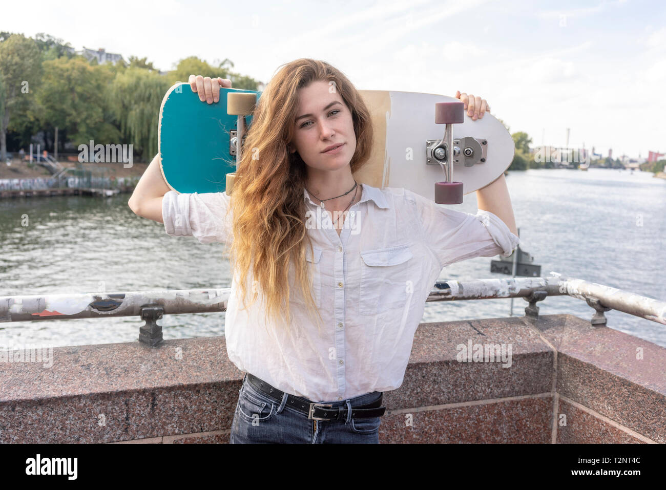 Mujer joven con el monopatín en el hombro, en el fondo del río, Berlín, Alemania Foto de stock
