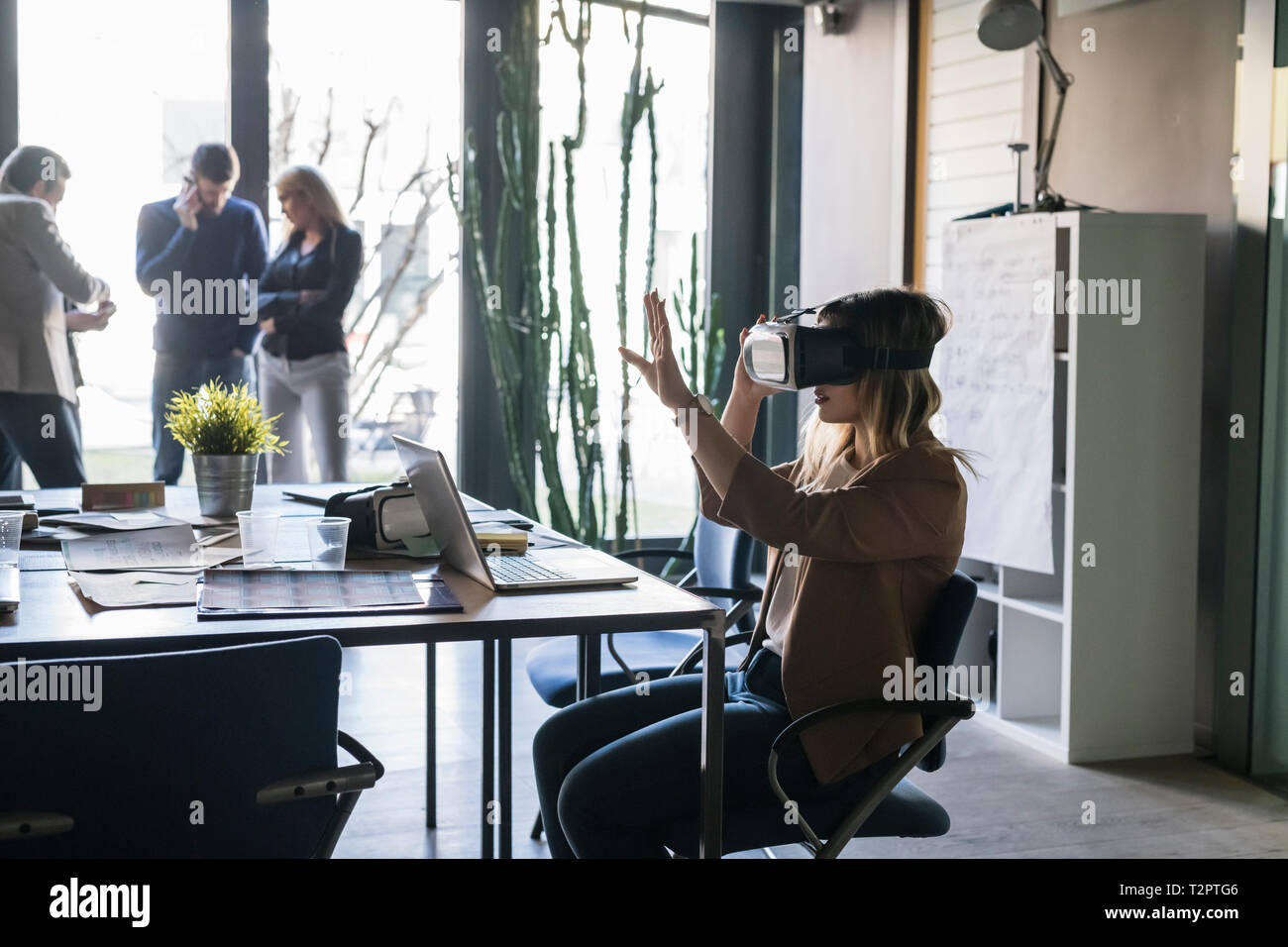 La empresaria con casco de realidad virtual en una oficina, colegas en segundo plano. Foto de stock