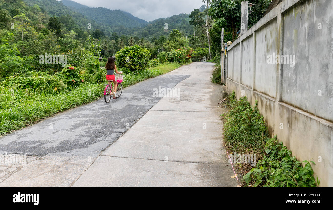 Chica en calzones rojos montando bicicleta junto a la pared en la carretera  en el pueblo en el bosque de lluvia Fotografía de stock - Alamy