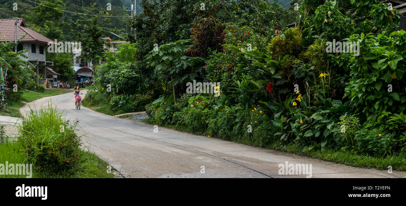 Chica en calzones rojos montando bicicleta en la carretera en el pueblo en el bosque de lluvia Foto de stock