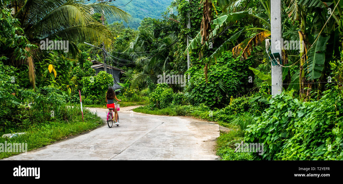 Chica en calzones rojos montando bicicleta en la carretera en el pueblo en el bosque de lluvia Foto de stock