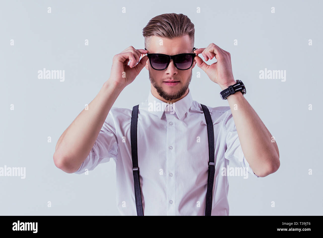 Apuesto y Elegante hombre en ropa clásica vistiendo gafas de sol mientras está pie contra un fondo gris. elegante con camisa blanca y suspender Fotografía de stock -