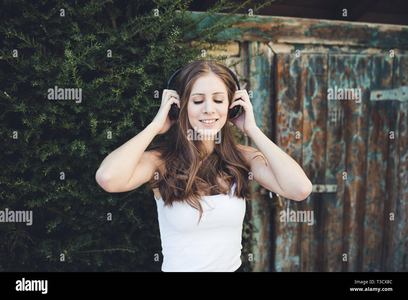 Mujer joven en una camiseta blanca sonriente con los ojos cerrados y escuchar música a través de auriculares al aire libre, campo. Foto de stock