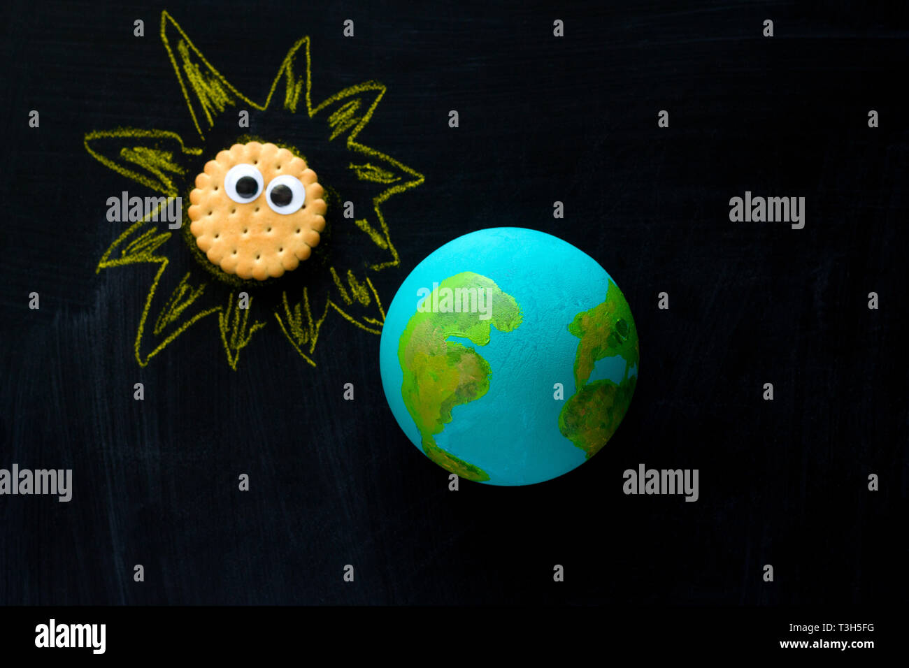 Vista superior del modelo artesanal del planeta Tierra y cookie gracioso Sunwith googly ojos en la pizarra , el espacio y la astronomía concepto Foto de stock