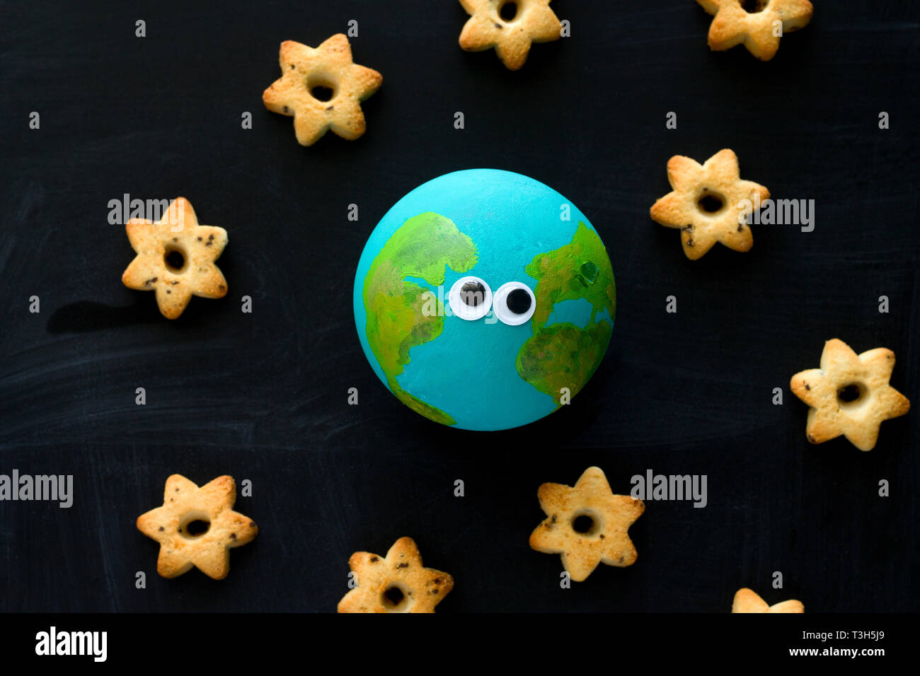 Vista superior del modelo artesanal del planeta Tierra con gracioso googly ojos y galletas en forma de estrellas en la pizarra , el espacio y la astronomía concepto Foto de stock