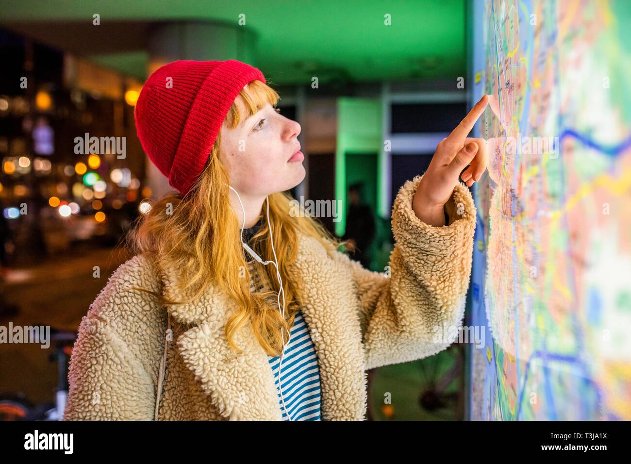 Niña, adolescente, con una gorra roja y los auriculares en el oído de pie delante de un mapa de la ciudad en la noche, Colonia, Renania del Norte-Westfalia, Alemania Foto de stock