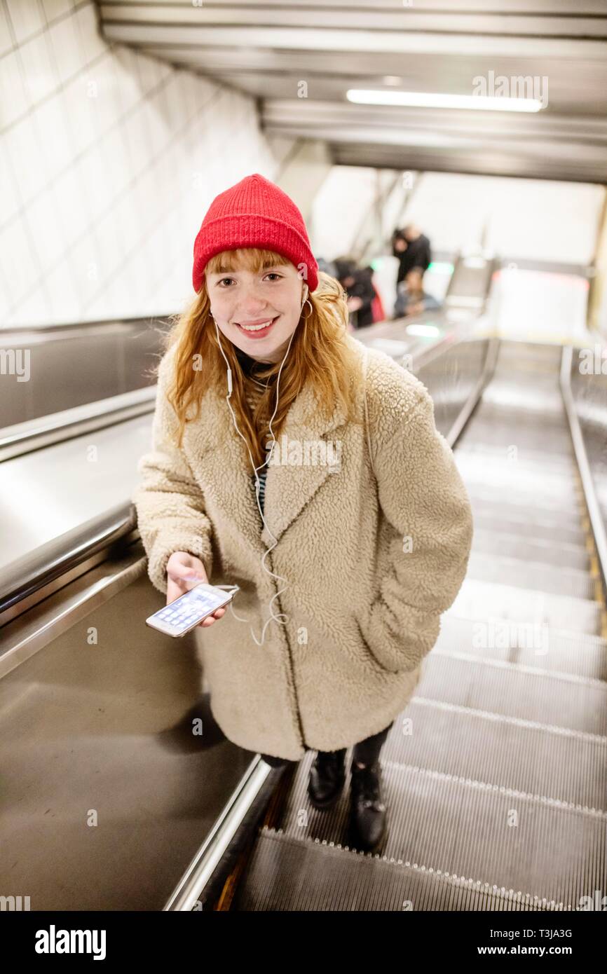 Niña, adolescente, con el smartphone en la mano y los auriculares en el oído en la escalera mecánica de una estación de metro, Colonia, Renania del Norte-Westfalia, Alemania Foto de stock