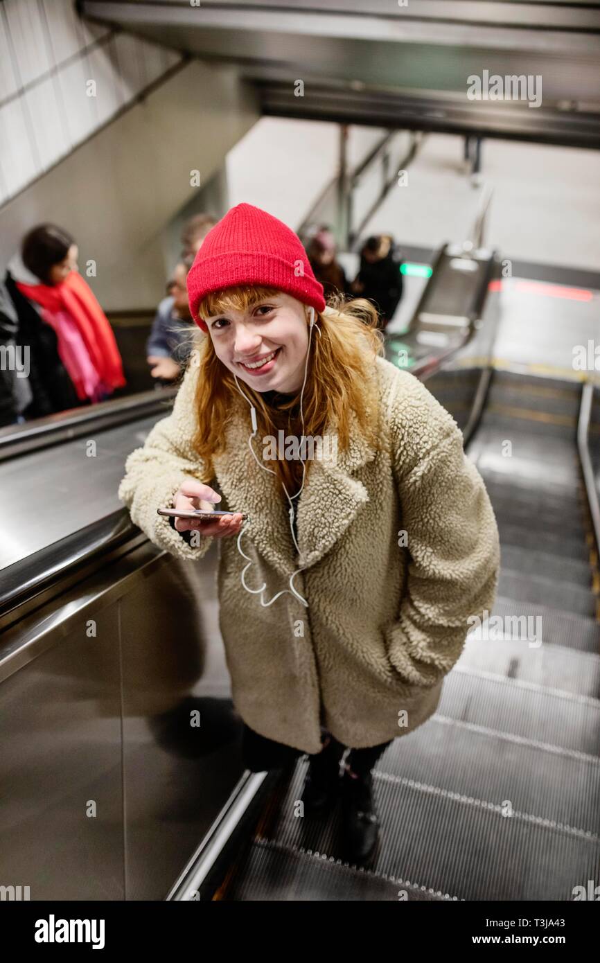 Niña, adolescente, con el smartphone en la mano y los auriculares en el oído en la escalera mecánica de una estación de metro, Colonia, Renania del Norte-Westfalia, Alemania Foto de stock