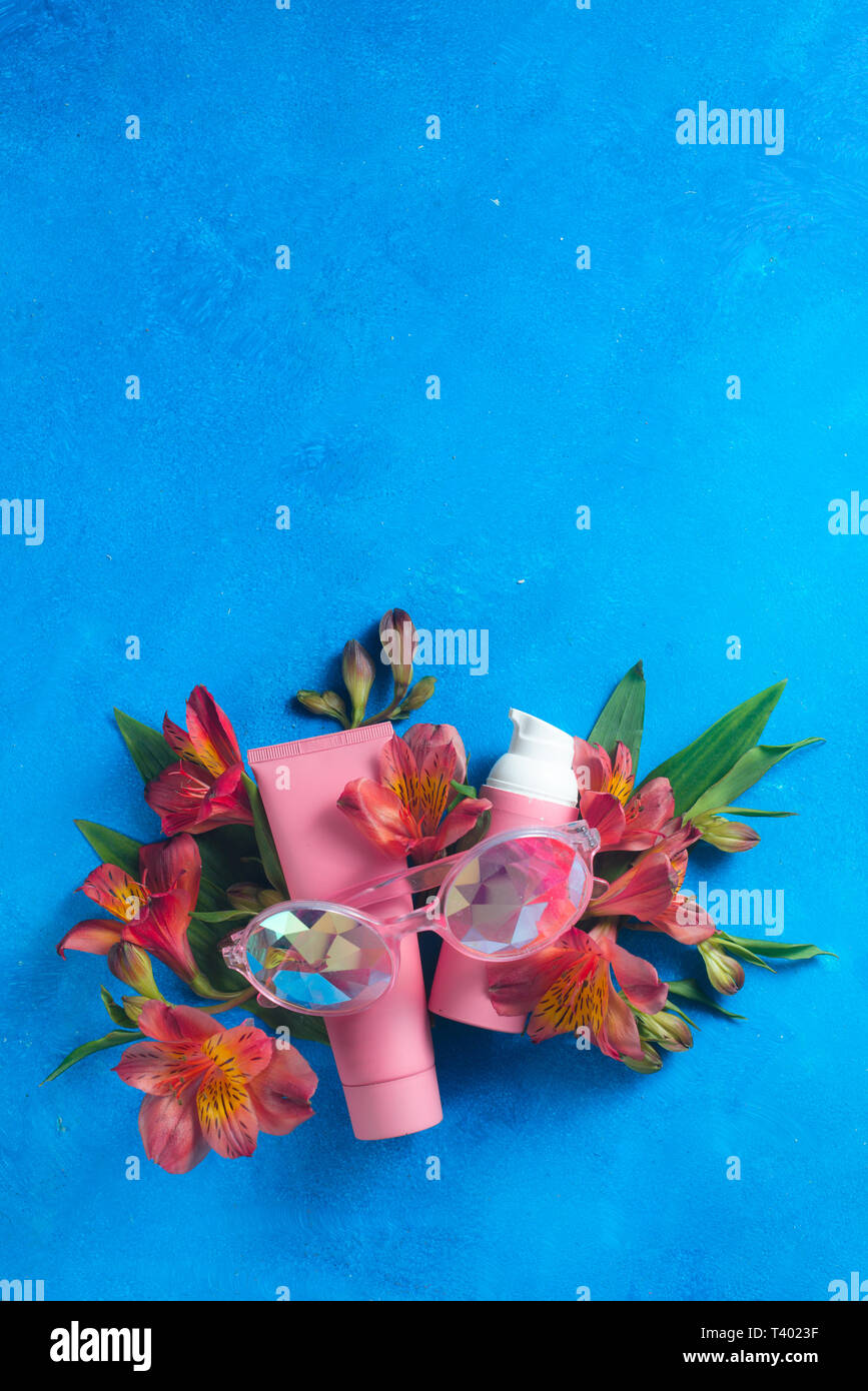 Gafas de sol y el cuidado de la piel cosméticos en la paleta de color rosa sobre un fondo azul con muelle de Alstroemeria flores. Sunscreen laicos plana con espacio de copia. Foto de stock